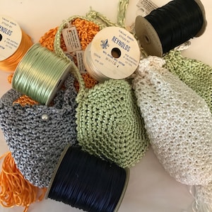 May include: A close-up of a collection of spools of yarn in various colors, including orange, green, white, and black. The yarn is arranged on a white surface, along with several crocheted bags in different colors and textures.