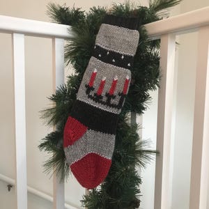May include: A gray and black knitted Christmas stocking with a red and white candle design. The stocking is hanging from a green garland.
