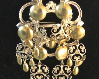 May include: A detailed vintage brooch with silver-tone filigree and gold-tone elements. The brooch has multiple gold spheres and teardrop dangles, creating a layered effect. The metal brooch is set against a dark background. The design is intricate and decorative.