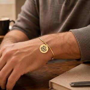 May include: A gold-toned metal bangle bracelet featuring a circular charm engraved with the text "BEST DAD EVER" flanked by two small leaf motifs. The bracelet has a thin, polished band and a simple hook closure mechanism on the charm.