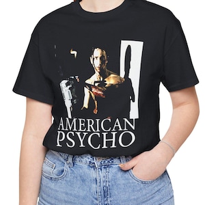Puede incluir: Camiseta negra con un gráfico de la película "American Psycho". El diseño muestra a un hombre sosteniendo un arma, con el título de la película en letras blancas debajo. La persona lleva vaqueros azules.