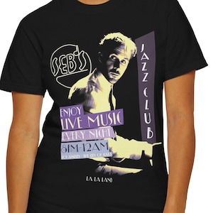 Puede incluir: Camiseta negra con un diseño gráfico retro para el Seb's Jazz Club. El diseño incluye un retrato de un hombre, el nombre del club y el texto "Enjoy Live Music Every Night 6PM 12AM" en morado y blanco. La camiseta la lleva una persona.