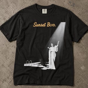 Könnte beinhalten: Ein schwarzes Kurzarm-T-Shirt mit einem Grafikdruck in Weiß und Beige. Das Design zeigt den Text Sunset Blvd. in Beige sowie eine weiße Illustration einer Person an einem Swimmingpool unter einem Scheinwerferlicht auf der Vorderseite.
