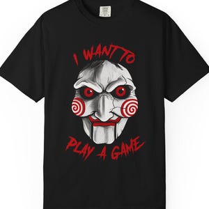 May include: Black t-shirt with a graphic of a creepy puppet face with red eyes and spiral cheeks. The text "I WANT TO PLAY A GAME" is printed in red above and below the face. The shirt is a short-sleeved crew neck.