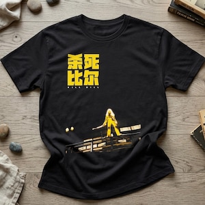 May include: A black short-sleeved t-shirt featuring a yellow graphic print. The design includes Chinese characters above the text "KILL BILL" and a central image of a figure in a yellow jumpsuit holding a sword on a platform structure.