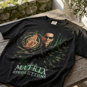 May include: A black short-sleeve t-shirt featuring a graphic print of Neo from The Matrix Revolutions. The design shows the character with sunglasses, a hand raised, and digital green code rain. The text Matrix Revolutions is printed below.