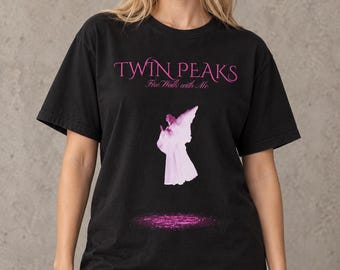 May include: A black short-sleeve t-shirt featuring the pink text TWIN PEAKS Fire Walk with Me. Below the text is a graphic of a light pink angel figure hovering above a glowing pink circular patch of light on the fabric of the shirt.
