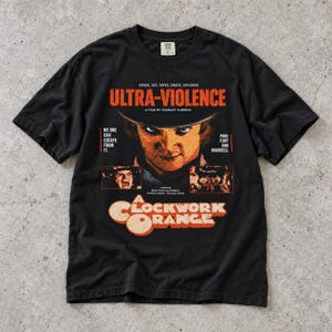 Peut inclure: Un t-shirt noir à manches courtes avec une impression graphique du film A Clockwork Orange. Le motif comprend le texte Ultra-Violence, un portrait d'un personnage portant un chapeau melon et des vignettes de scènes avec des lettres orange et blanches.