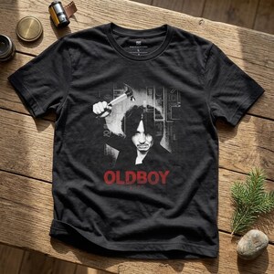 Pode incluir: Uma t-shirt preta de manga curta com uma impressão gráfica de uma pessoa a segurar um martelo acima da cabeça. O design inclui o texto "OLDBOY" em letras maiúsculas vermelhas abaixo da figura, sobre um fundo de linhas verticais.