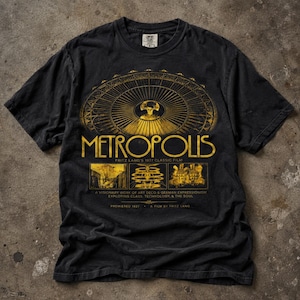 Puede incluir: Una camiseta negra de manga corta con un estampado gráfico dorado de la película Metropolis de 1927. El diseño incluye un gran motivo arquitectónico circular, el título Metropolis, tres escenas rectangulares pequeñas y texto sobre el filme.