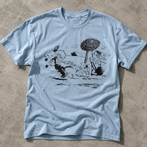 May include: A light blue short-sleeve t-shirt featuring a black ink-style graphic print. The illustration depicts a cartoon character running with the text "POW" and "ZIP" nearby, alongside a tree and a figure in a uniform holding a baton.