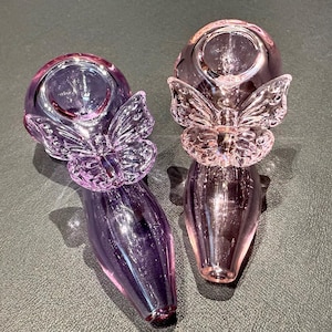 May include: Two glass pipes, one purple and one pink, each featuring a butterfly design. The pipes have a rounded bowl and a slender stem. The butterfly is positioned near the bowl.