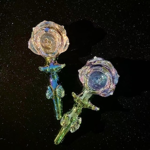 May include: Two iridescent glass rose-shaped objects with a stem and leaf design. The glass has a swirling pattern of colours including blue, green, and gold. The objects are on a black surface.