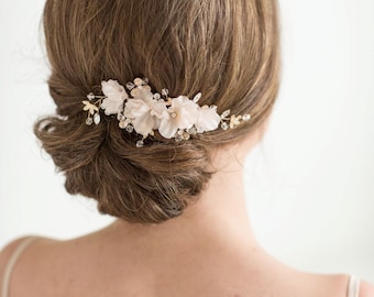 Puede incluir: Un peine para el cabello delicado con flores de tela blanca y detalles dorados. El peine está adornado con pequeños cristales y cuentas.