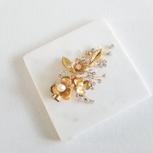 May include: Gold hair comb with a floral design featuring a cluster of small flowers, leaves, and rhinestones. The comb has a pearl accent.
