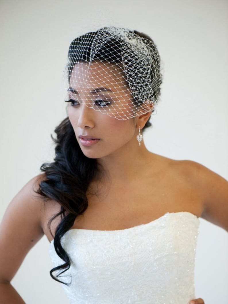 May include: A bride with long dark hair styled in a side braid, wearing a white strapless wedding dress and a white birdcage veil.