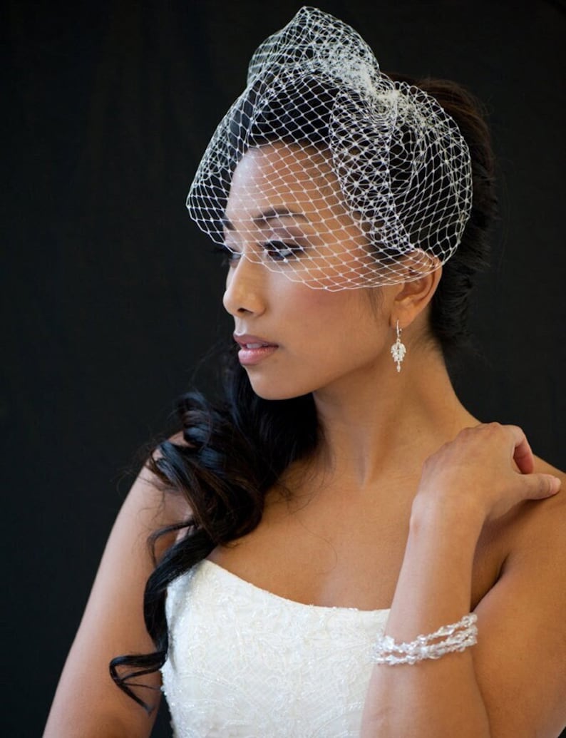 May include: A bride wearing a white strapless wedding dress with a white birdcage veil. She has long dark hair styled in a side braid and is wearing a crystal bracelet and earrings.