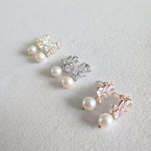 May include: Three pairs of elegant stud earrings. Each earring features a cluster of clear crystals and a dangling pearl. The earrings are in gold, silver, and rose gold tones, set against a white background.