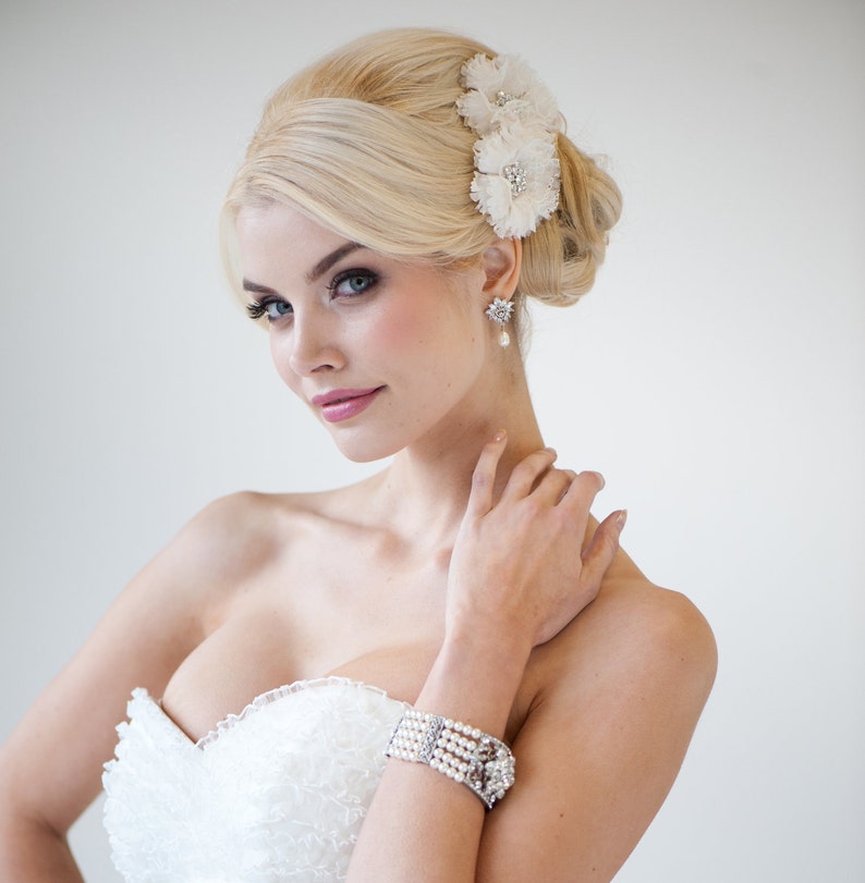 May include: A bride wearing a white strapless wedding dress, a pearl and diamond bracelet, and flower hair accessories. The bride also wears a pair of star-shaped earrings with a pearl drop. The bride has blonde hair and is looking at the camera.