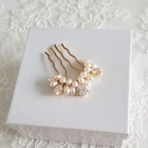 Puede incluir: Un peine de pelo dorado con una fila de perlas y dos diamantes de imitaci&oacute;n transparentes. El peine est&aacute; sobre una superficie blanca.