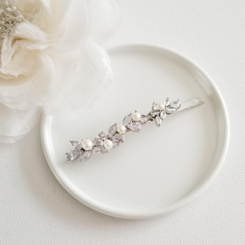 May include: A silver-toned hair barrette with a floral design. The barrette features clear, sparkling crystals and faux pearls. The barrette is on a white plate, with a white flower in the background.