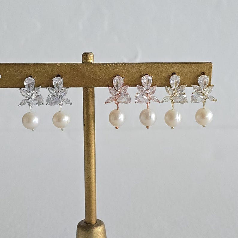 May include: A display of six earrings, each featuring a cluster of clear, sparkling crystals in a floral design. Below the crystals, a single, round, white pearl dangles. The earrings are presented on a gold-colored stand against a white background.