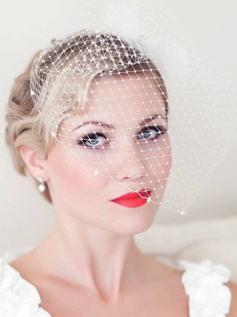 May include: A bride with blonde hair wearing a white veil with pearl accents and a red lipstick.