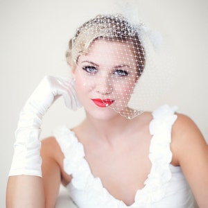 May include: A bride wearing a white dress with a white veil and a white glove. She has red lipstick on and is looking at the camera.
