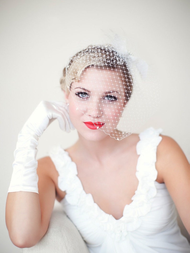 May include: A bride wearing a white dress with a white veil and a white glove. She has red lipstick on and is looking at the camera.
