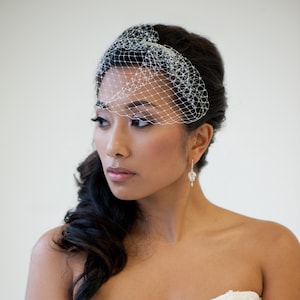 Puede incluir: Una novia con el pelo oscuro peinado en una coleta lateral lleva un velo blanco con un delicado patrón de red. Lleva un vestido de novia blanco sin tirantes y pequeños pendientes de cristal.
