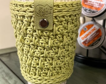 May include: A green crocheted coffee cup cosy with a brown leather strap and a small metal buckle. The cosy is on a white paper coffee cup with a grey and white pattern. The cup is sitting on a wooden surface.