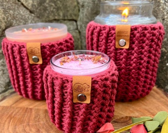 May include: Three red crocheted candle holders with leather labels. The candle holders are on a wooden surface with a bouquet of red flowers.