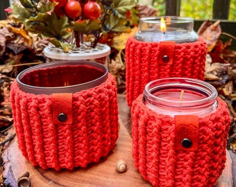 Könnte beinhalten: Drei Glaskerzenbehälter mit roten Häkelüberzügen. Die Überzüge haben einen braunen Lederriemen mit einem schwarzen Knopf. Die Kerzen stehen auf einer Holzoberfläche mit Herbstblättern.