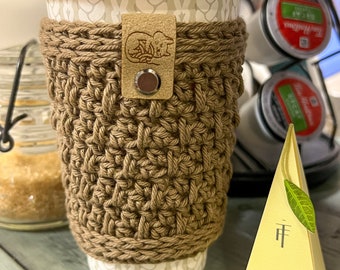 May include: A brown crocheted coffee cup cosy with a leather patch and a metal button. The cosy is on a white coffee cup with a white and grey pattern. A box of Jasmine Green tea is in the background.