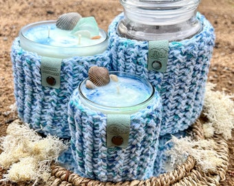 Könnte beinhalten: Drei Kerzen in Glasbehältern mit blauen und weißen Häkelüberzügen. Die Kerzen stehen auf einem geflochtenen Tablett mit getrocknetem Moos.