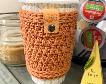 May include: A crocheted coffee cup cosy in a burnt orange colour with a leather strap and a small metal button. The cosy is on a white takeaway coffee cup. A box of chamomile citron herbal tea is in the background.