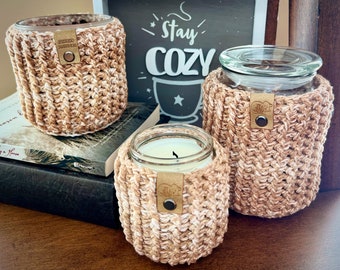 Puede incluir: Tres tarros de velas de crochet en beige y blanco con una etiqueta de cuero marrón. Los tarros están sobre una superficie de madera con un letrero que dice "Stay Cozy".