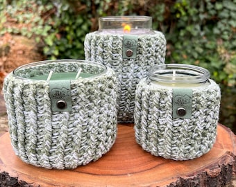 Könnte beinhalten: Drei Glas-Kerzenbehälter mit grünen und weißen Häkelüberzügen. Jeder Überzug hat ein Leder-Etikett mit dem geprägten Wort "Snuggle".