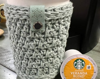 May include: A grey crocheted coffee cup cosy with a leather strap and snowflake design. A Starbucks Veranda Blend K-Cup pod is next to the cosy. A yellow packet of Stevia in the Raw zero calorie sweetener is in the foreground.