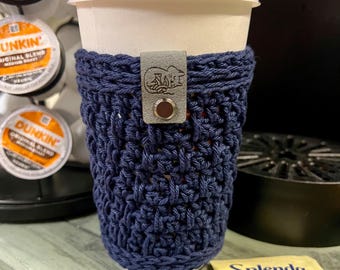 May include: A navy blue crocheted coffee cup cosy with a leather strap and a small metal button. The cosy is on a white paper coffee cup.