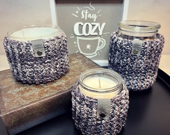 Puede incluir: Tres tarros de vidrio para velas con velas blancas en su interior. Cada tarro está envuelto en un 'cozy' de punto gris y blanco. El letrero detrás de los tarros dice "Stay Cozy".
