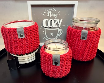 Puede incluir: Tres portavelas de crochet rojo con una etiqueta gris. Los portavelas están sobre una superficie negra. El letrero en el fondo dice "Mantente acogedor".