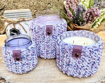 Könnte beinhalten: Drei violette und weiße gehäkelte Kerzenhalter mit Leder-Etiketten. Die Kerzen befinden sich in Glasbehältern und sind angezündet. Die Kerzenhalter stehen auf einer Holzoberfläche.