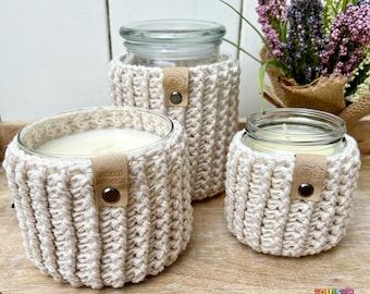 Könnte beinhalten: Drei Glas-Kerzenbehälter mit weißen Kerzen darin. Jede Dose ist in eine gehäkelte cremefarbene Hülle mit einem braunen Leder-Etikett mit der Aufschrift "SQUISHY PRODUCTS" eingewickelt.