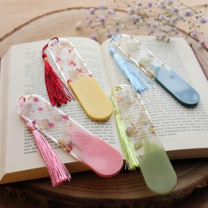 Peut inclure: Cinq marque-pages en résine colorée avec des pompons reposent sur un livre ouvert. Les marque-pages présentent des fleurs séchées et des paillettes dorées incrustées. Couleurs : rose, jaune, vert, bleu et résine transparente.