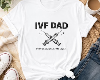 May include: White t-shirt with the text "IVF DAD" above a graphic of two crossed syringes. Below the graphic, the text reads "PROFESSIONAL SHOT GIVER". The shirt is styled with a knot at the bottom.