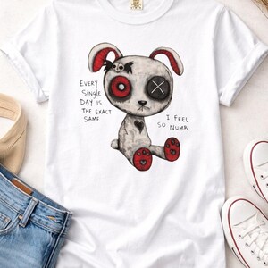 May include: White t-shirt featuring a distressed illustration of a bunny-like creature with one red eye and one X eye. The text reads "Every single day is the exact same. I feel so numb."