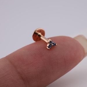 May include: A rose gold lip piercing stud with a flat disc backing and a post. The stud features a cluster of three small, dark blue gemstones in a triangular arrangement. The jewelry is shown against a neutral background.