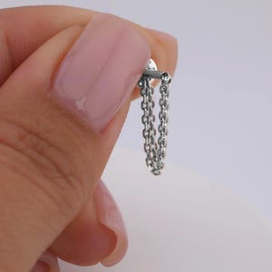 May include: A silver chain earring with a small stud and a dangling chain. The earring is held in a hand against a white background. The chain is made of small, interconnected links.
