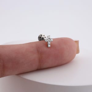 May include: A small, silver-colored cross earring with multiple clear, sparkling stones. The cross is the focal point, with a flat back and a post for piercing. The earring is shown on a finger against a white background.